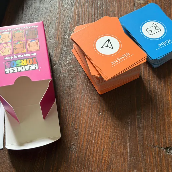 Headless Torsos pink box open on a table beside stacked orange ANSWER cards and blue INBOX cards
